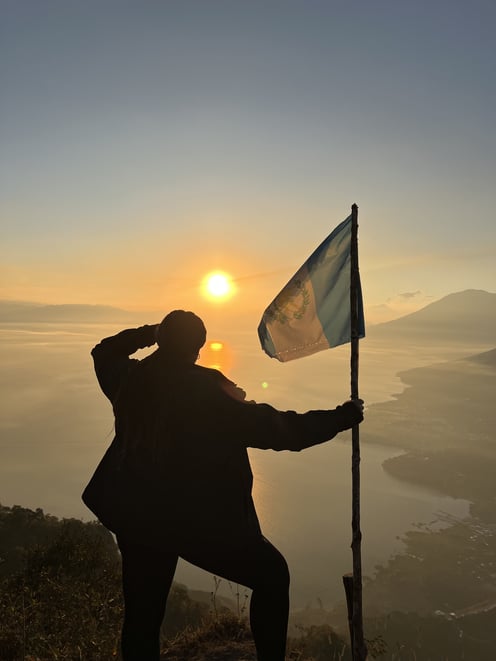 A photo of a woman, holding a Guatemalan flag, overlooking a shoreline, with the sun low on the horizon. 